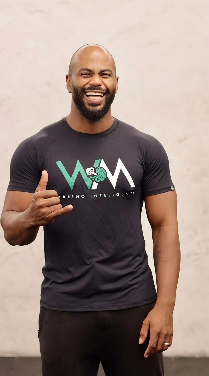 Will Mesquitta sorrindo, de camiseta preta com o logo WM, fazendo o gesto de hang loose com a mão.